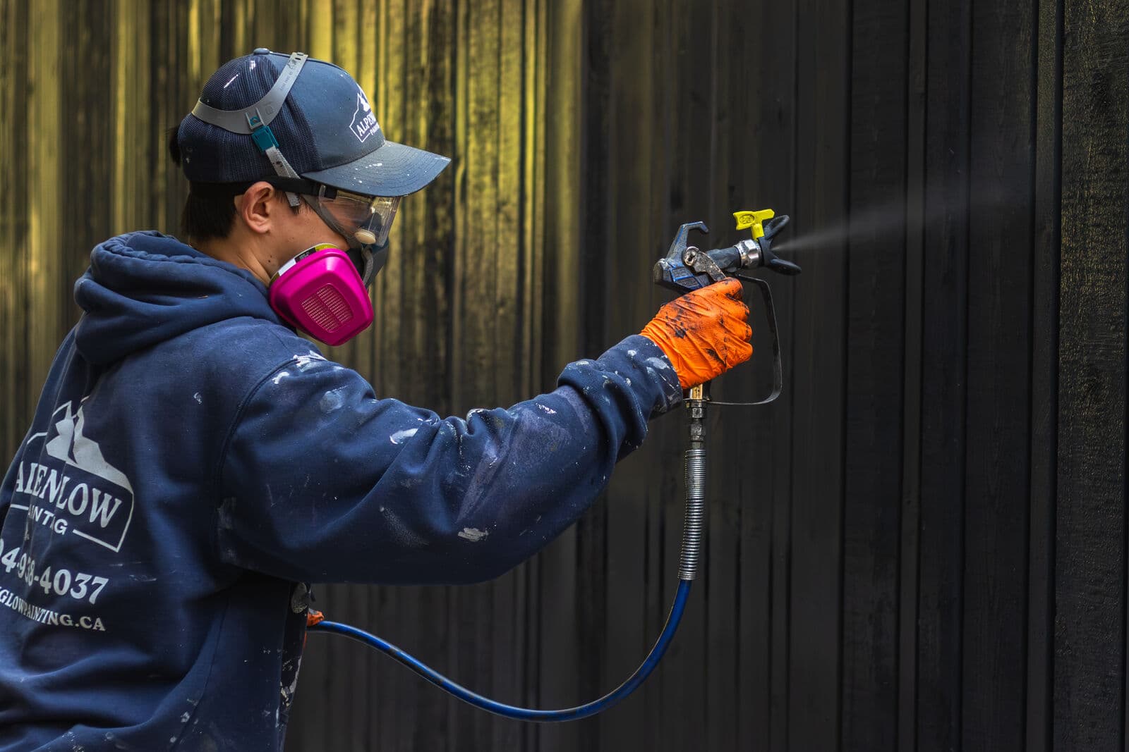 Side profile of a painter in a navy Alpenglow Painting hoodie, cap, pink respirator