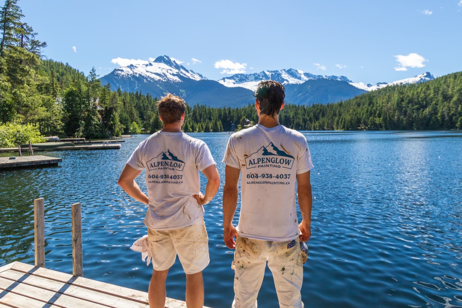 Two crew members in paint-spattered white Alpenglow Painting branded t-shirts stand on a wooden dock looking across a blue lake toward snow-capped