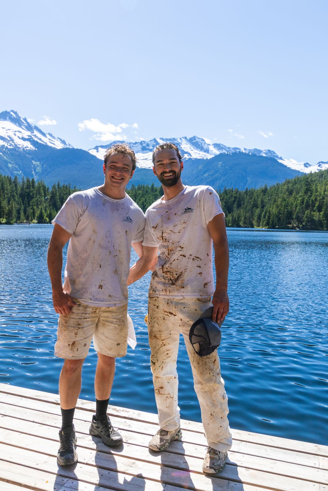 Two smiling Alpenglow Painting crew members in paint-splattered white logo t-shirts and whites stand full-length on a wooden dock with a blue lake