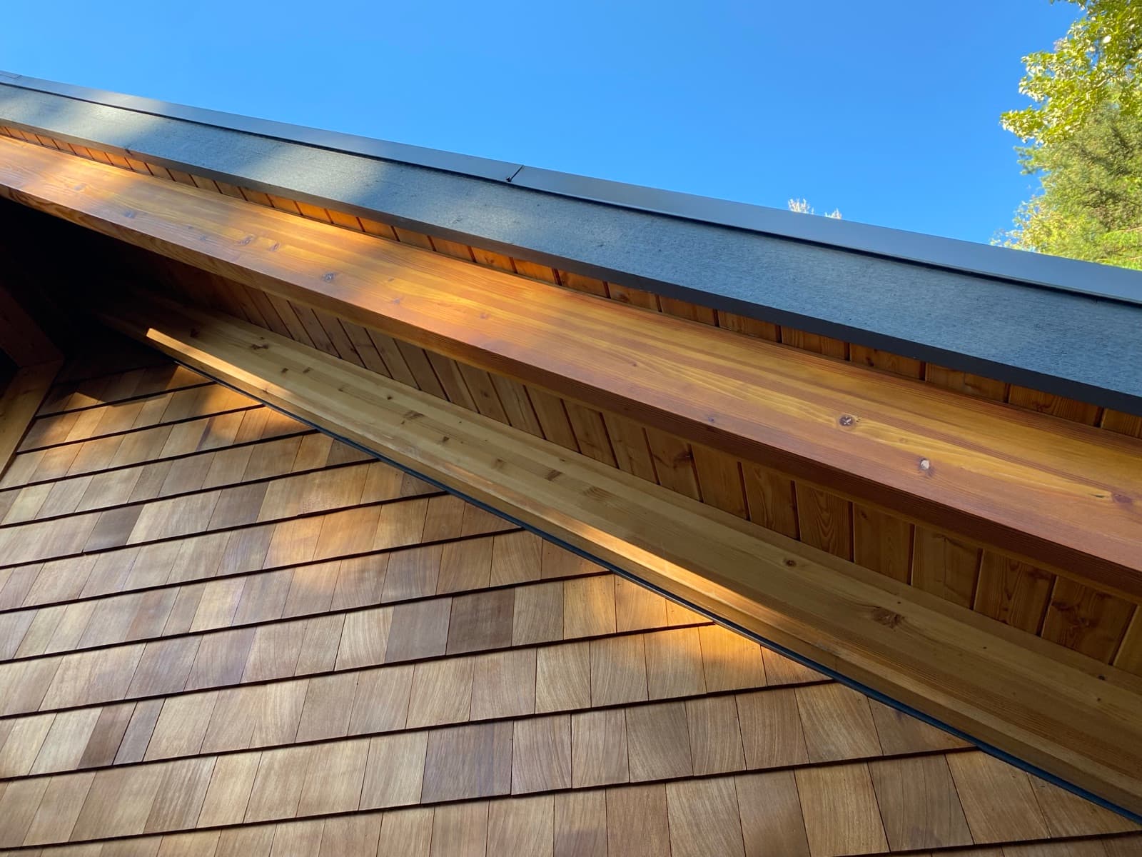 Low-angle close-up of freshly stained natural cedar shake siding and warm-toned tongue-and-groove soffit boards under a dark fascia against blue sky