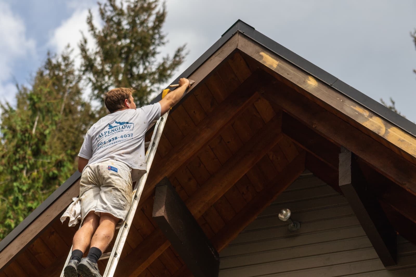 Landscape view of a painter in an Alpenglow Painting branded shirt on a ladder applying stain to the fascia board along a chalet's exposed cedar