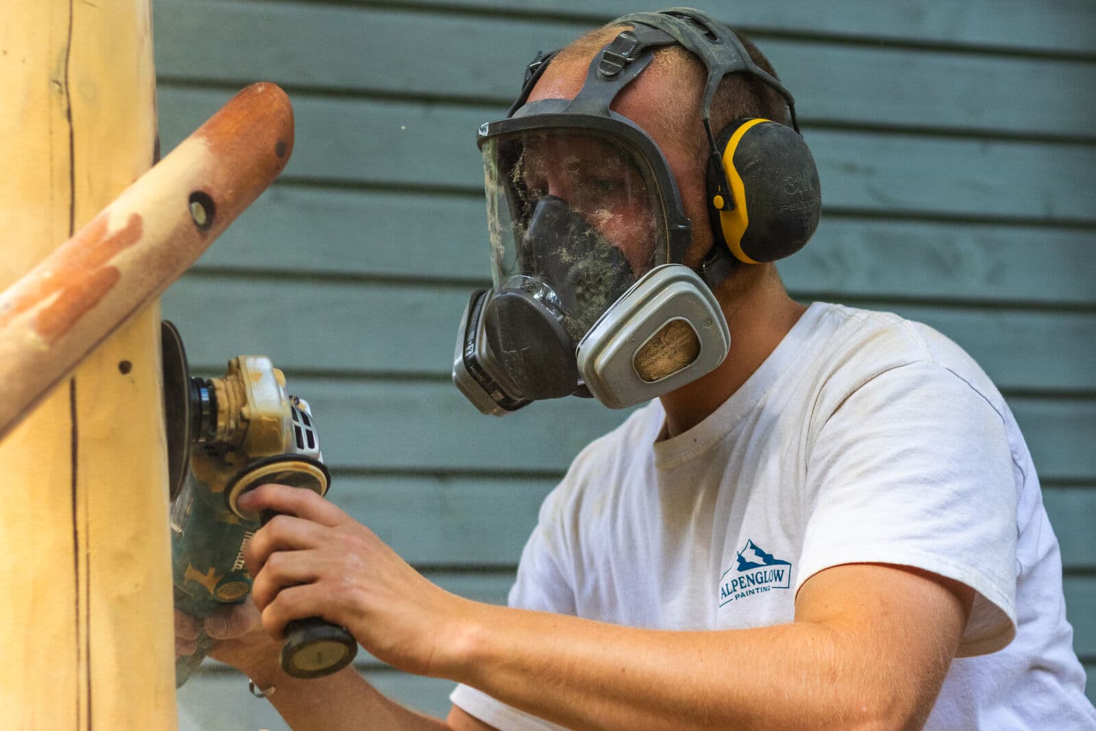 Slightly wider angle of the same respirator-clad worker gripping a Makita sander with both hands while stripping old finish from a log post against