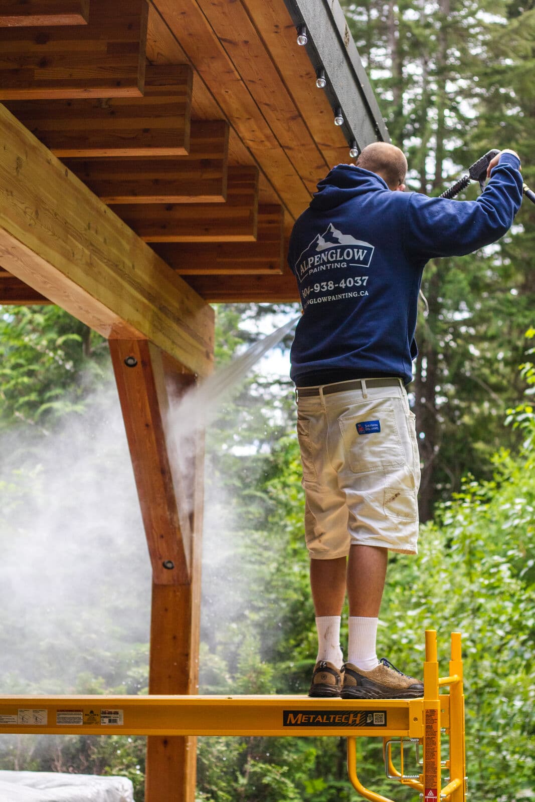 Alpenglow crew member in branded hoodie standing on yellow scaffolding pressure-washing exposed cedar soffit beams and timber posts before refinishing