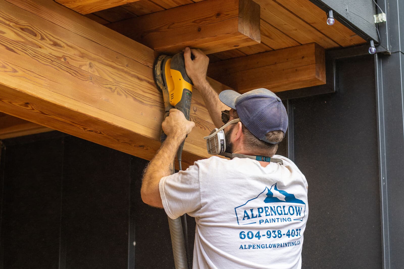 Painter sanding the underside of an exposed wood beam and tongue-and-groove soffit against black metal siding, sander partly hidden overhead