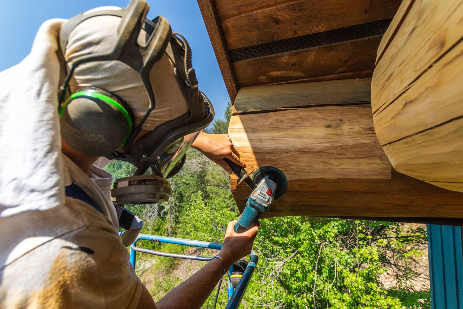 Worker in full-face respirator and hearing protection on blue scaffolding uses an angle grinder with sanding disc to strip weathered finish from a