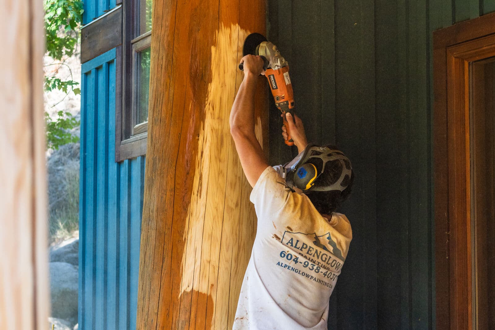 Crew member in Alpenglow Painting shirt reaches overhead with a Ridgid grinder to strip stain from a log porch post