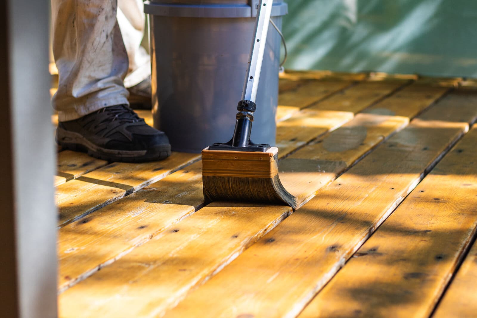 Low-angle close-up of a wide stain brush on a pole applying warm golden stain to sunlit cedar deck boards