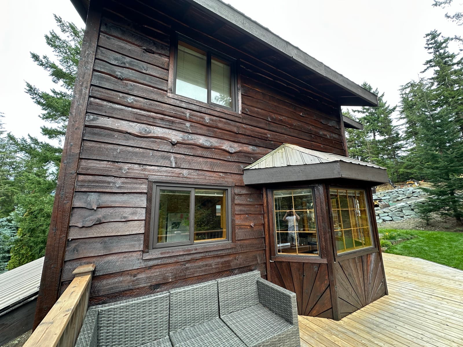 Two-storey home with rough-sawn board siding freshly stained dark espresso brown, metal-roofed bay window and new cedar deck with grey wicker seating