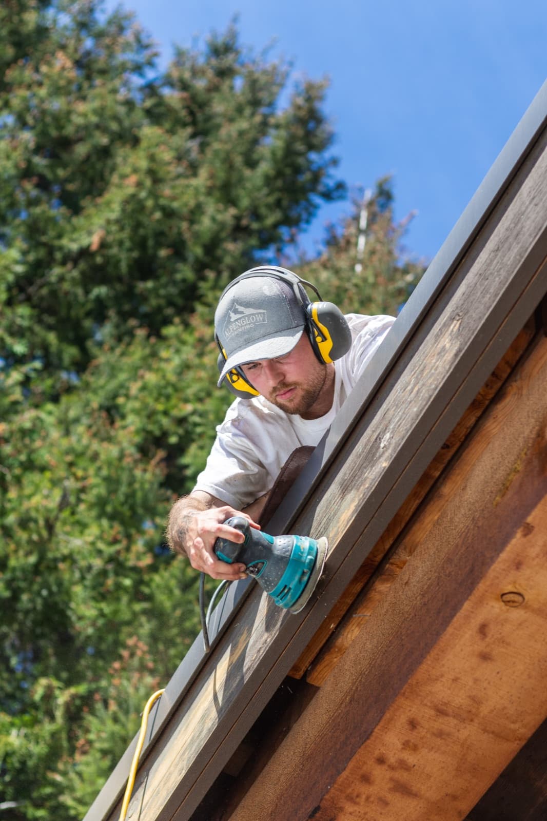 Vertical shot of a painter in an Alpenglow Painting cap and ear muffs sanding a grey weathered roof fascia board with a teal orbital sander