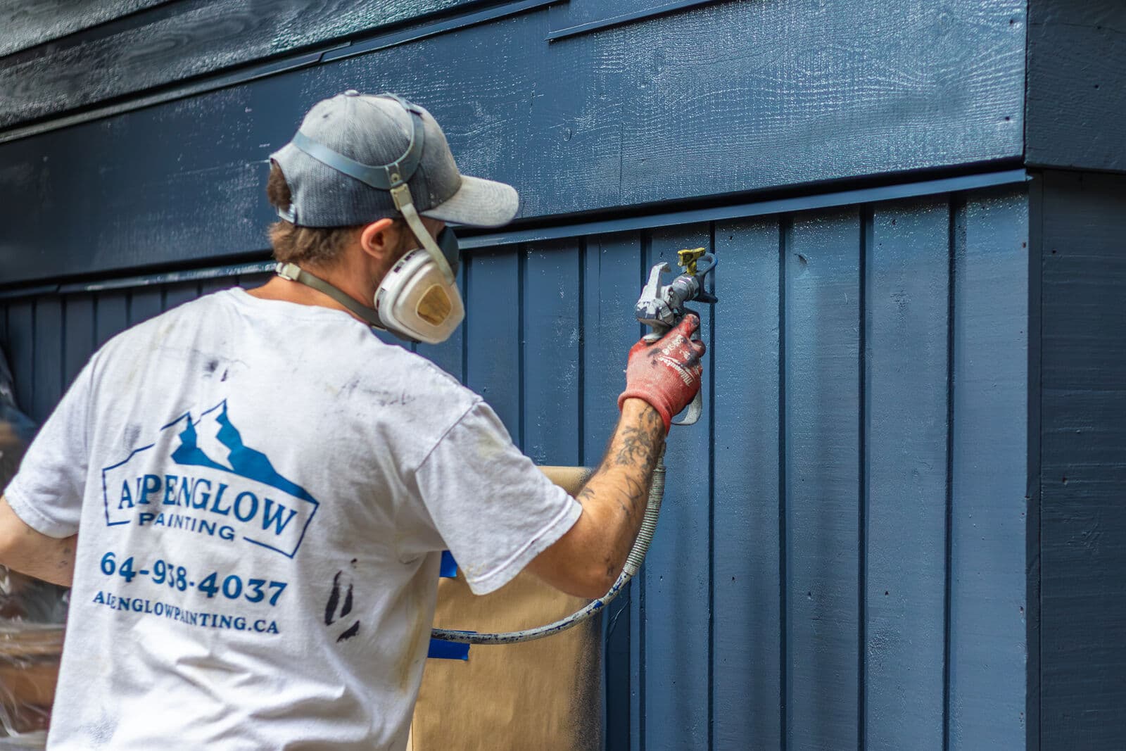 Painter in grey cap, respirator, and white Alpenglow Painting branded t-shirt applying a fresh wet coat of deep blue paint to vertical-panel siding