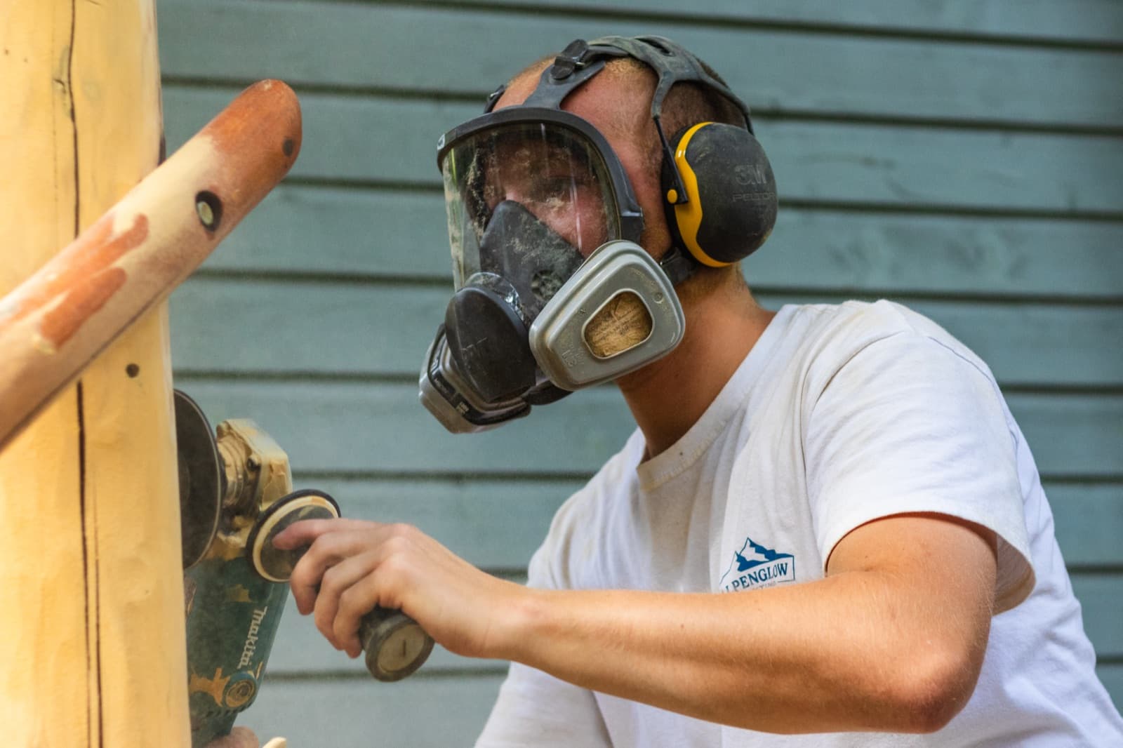 Close-up of a crew member wearing a full-face respirator and yellow 3M ear muffs sanding a log post back to bare wood with a Makita disc sander