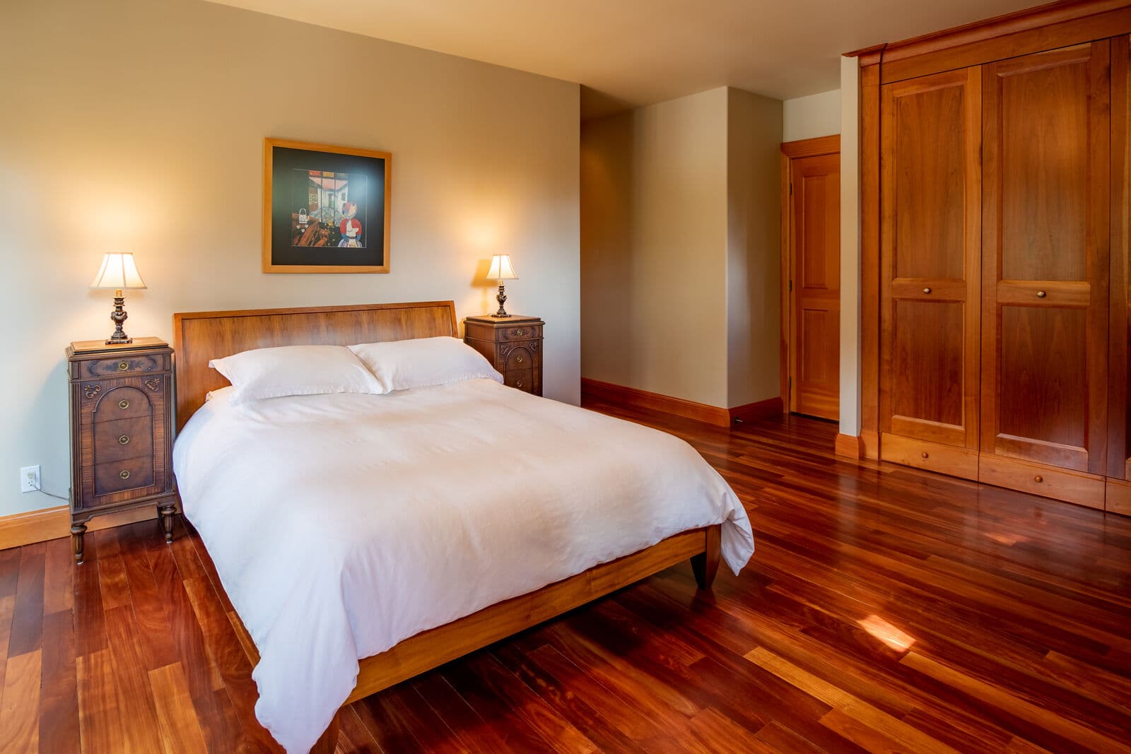 Alternate master bedroom angle showing greige painted walls, built-in cherry wardrobe doors, white bedding and polished exotic hardwood flooring