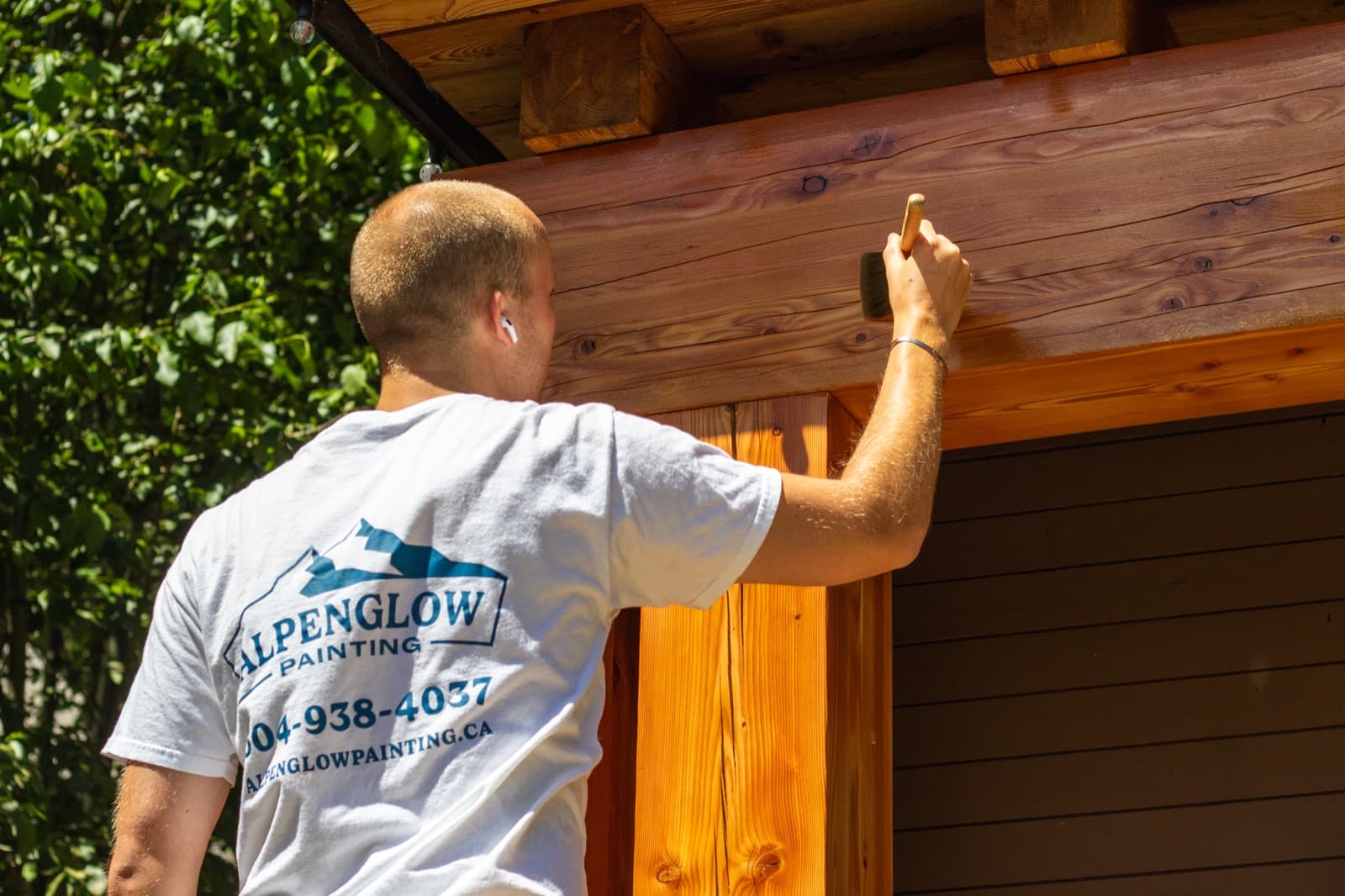Landscape view of an Alpenglow-shirted painter brush-applying stain along a large horizontal cedar header beam above dark brown horizontal siding
