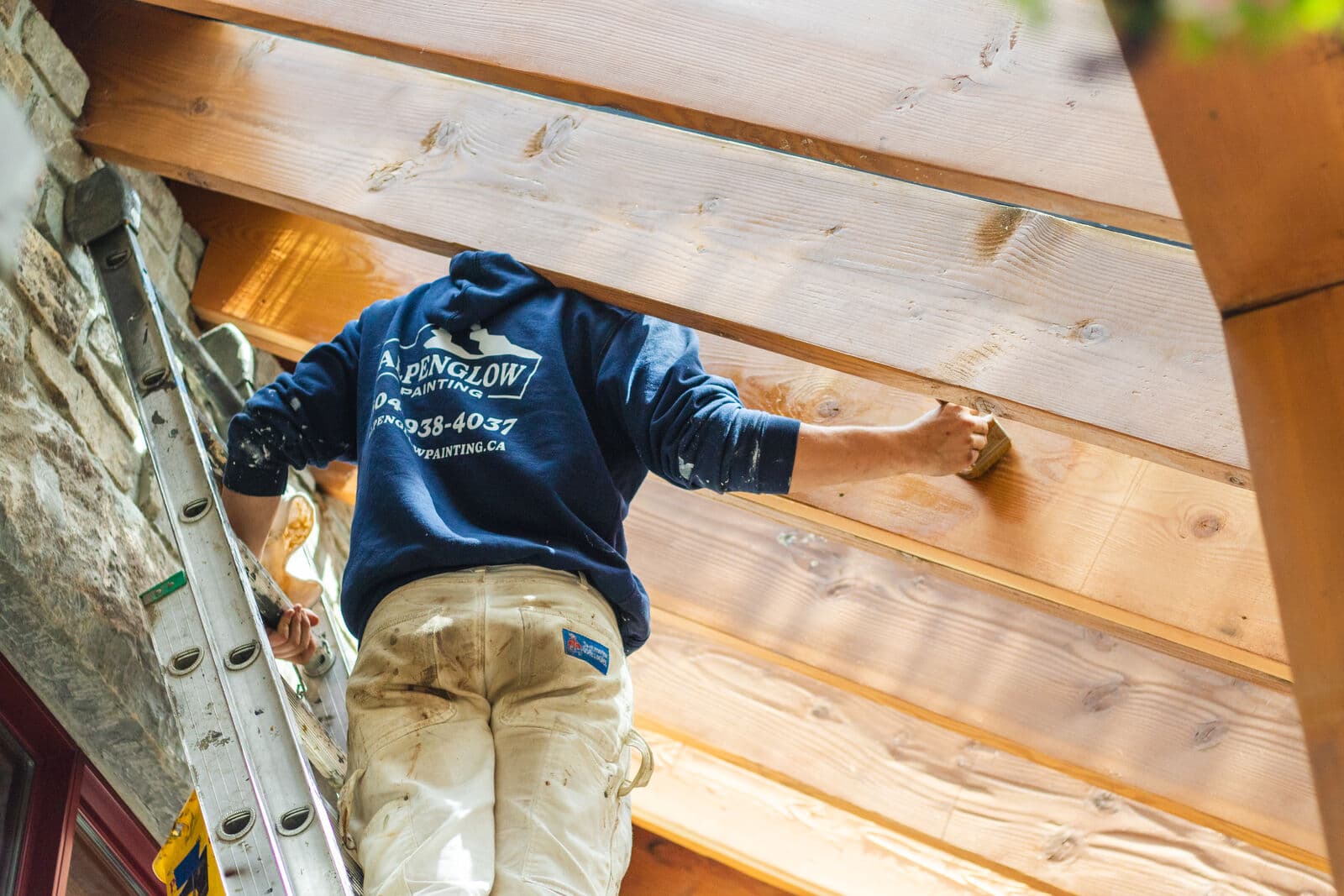 Painter on a ladder in an Alpenglow Painting branded navy hoodie hand-applies a whitewash stain to exposed tongue-and-groove soffit planks between