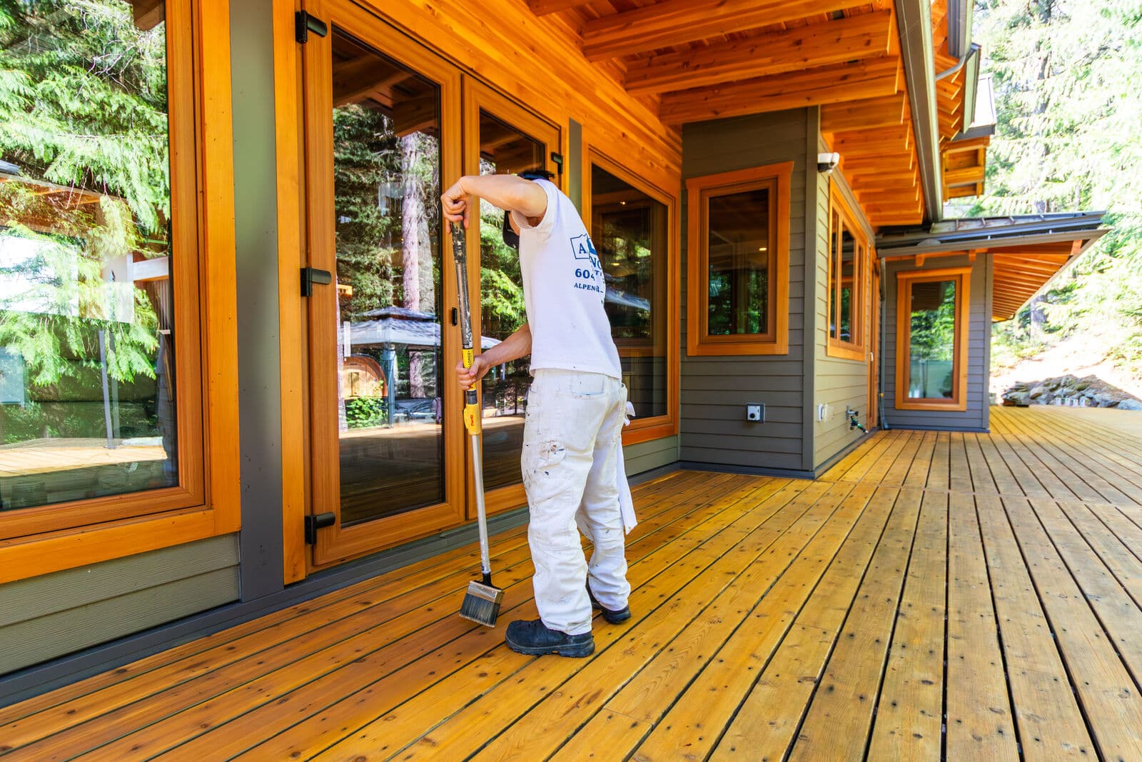 Wide-angle view of an Alpenglow Painting crew member in a branded white t-shirt and painter's whites using a Purdy pole brush to stain a large cedar