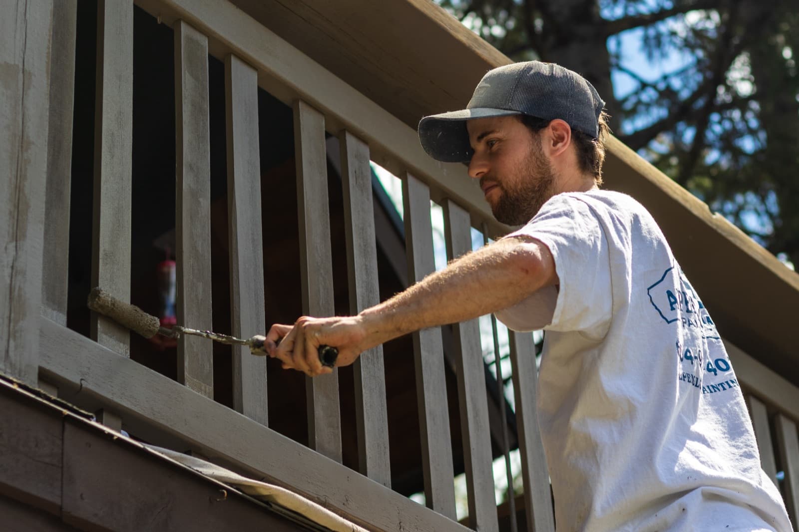 Bearded painter in a grey trucker cap applies grey-taupe coating to vertical deck railing balusters with a mini roller, face in profile