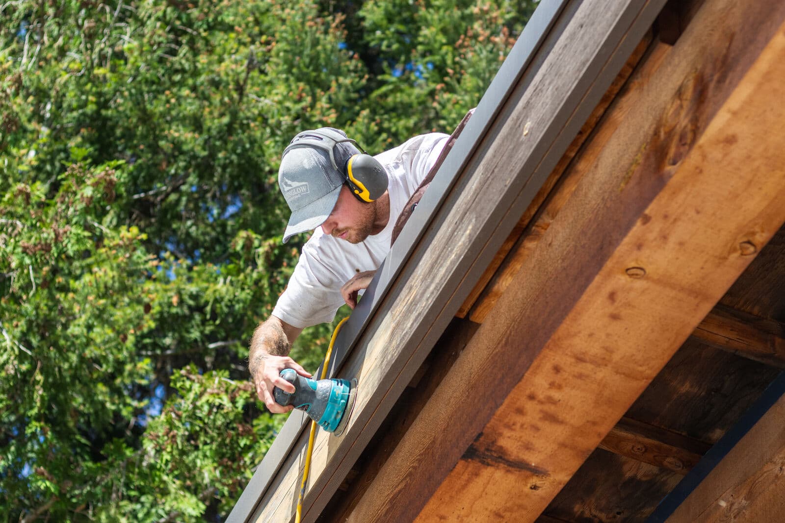 Wide diagonal shot of a painter in a grey Alpenglow cap and yellow ear muffs leaning over a roof edge to sand a weathered grey fascia board with a