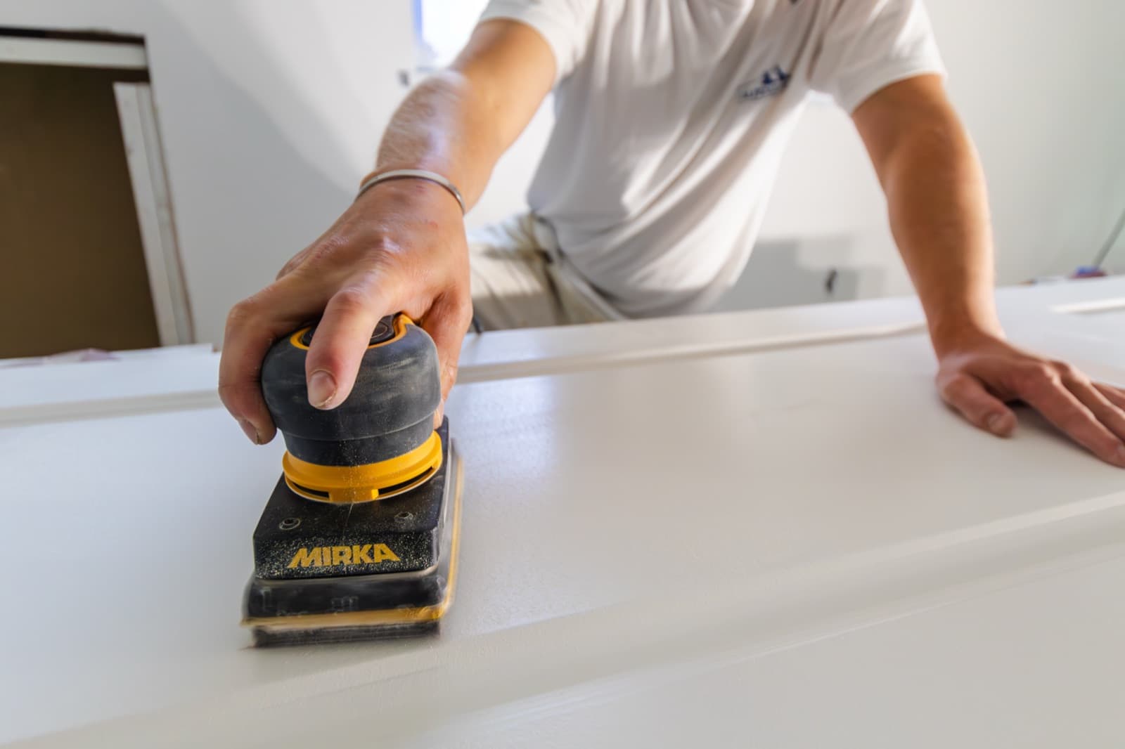 Painter in a white t-shirt with a small blue Alpenglow chest logo sanding a white door slab with a Mirka orbital sander