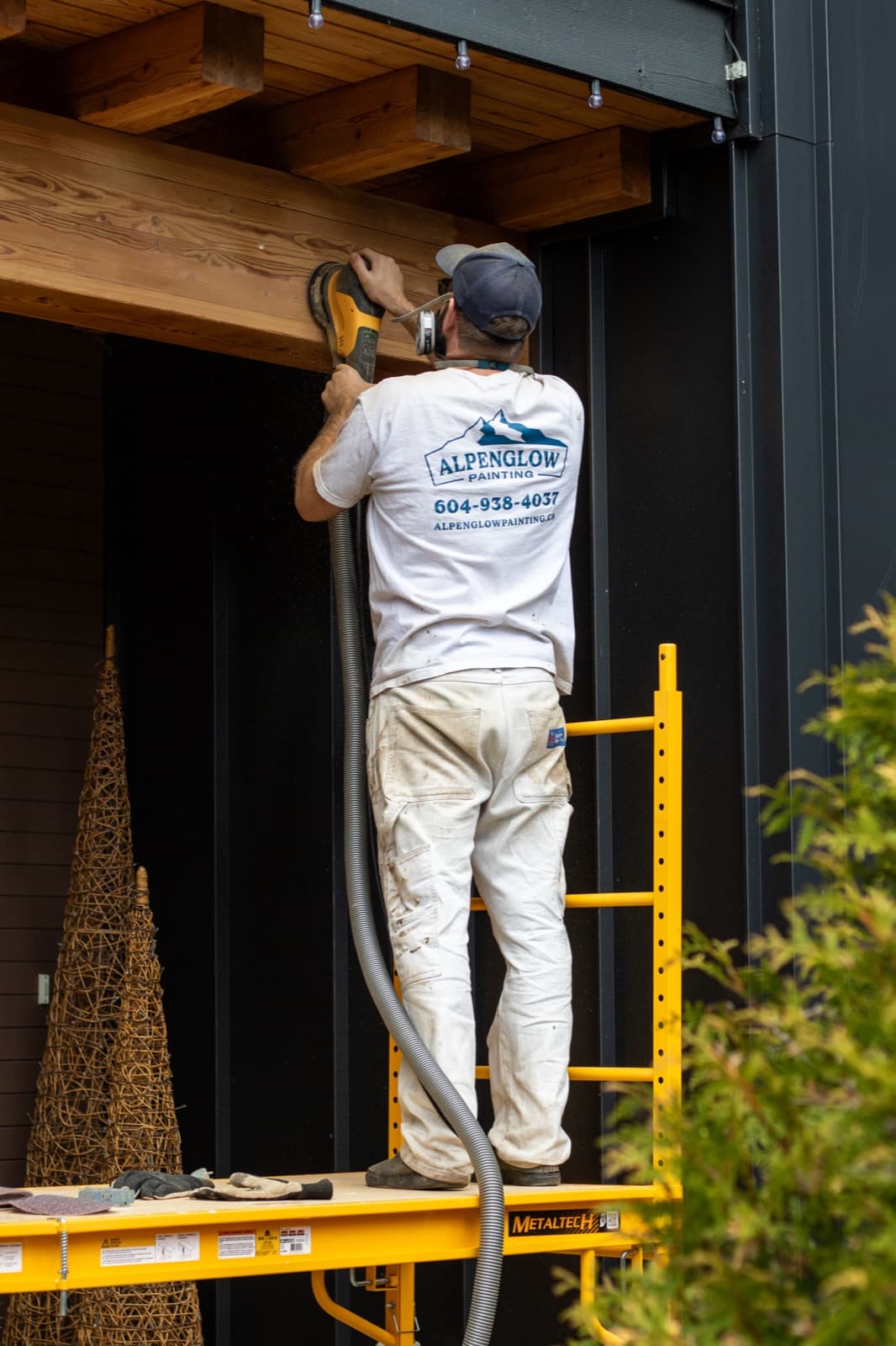 Full-body shot of a painter standing on a yellow Metaltech scaffold sanding a timber header beam beneath a wood soffit on a modern black-clad home